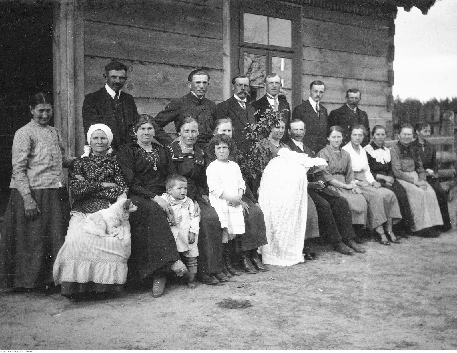 Village christening party of 1922 posing outside a wooden house, baby in a white gown