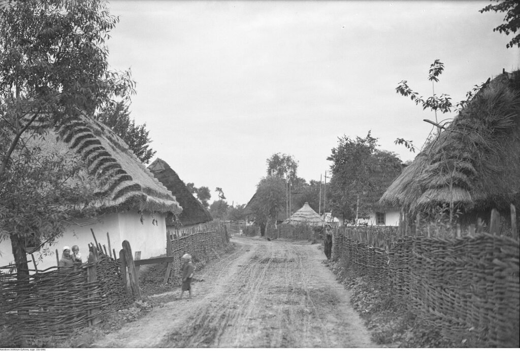 Dirt village street lined with thatched cottages and woven fences, 1934