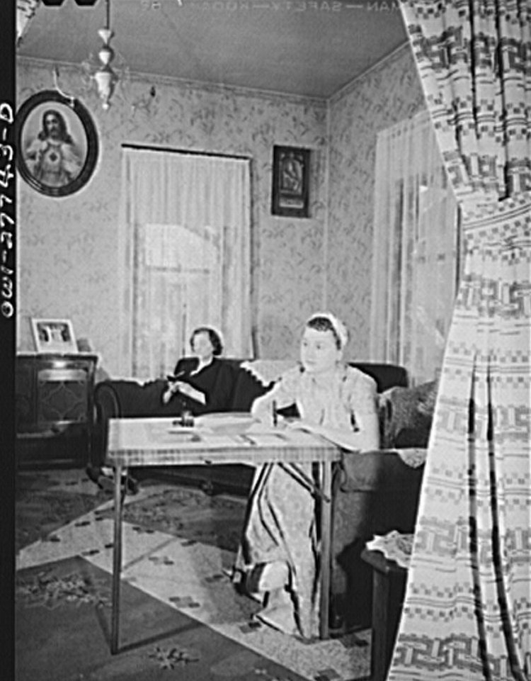 Young woman writing a letter at a table while an older woman reads a prayer book behind her, 1943